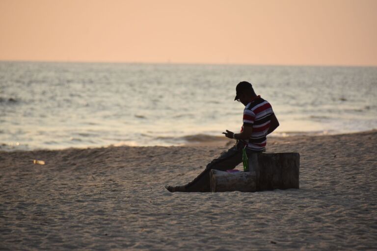 A man sits on a log at a beach, facing the sea, wearing a striped shirt and a cap, looking at a cell phone in his hand.